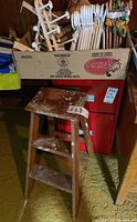 Wooden three step stool with paint splatters and a box of white plastic and wood hangers plus over-the-door hooks in the background