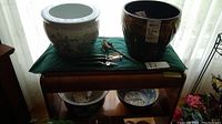 Overall view of two large ceramic plant pots placed on a table, with handheld gardening utensils placed beside them and two smaller planters/bowls visible below the table.
