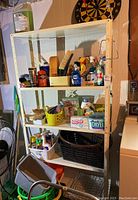 Full view of the white metal shelving unit with four adjustable shelves holding various household cleaning and storage items (not included in sale). Shelving is shown against a beige basement wall with a dartboard hung above the unit.