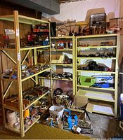 Three beige metal shelving units arranged in basement with various hand tools, small boxes, jars, cables, and hardware items stored on shelves