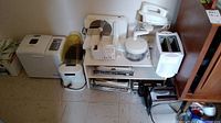Photo showing multiple small kitchen appliances including bread machine, toasters, stand mixer without bowl, popcorn maker, kitchen scales and meat slicer on floor against wall.