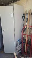 Tall closed gray metal storage cabinet with door closed and visible handle, with yard tools and hand truck beside it.