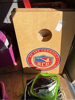 Front view of first American Cornhole wooden board with ACO logo and hole, with basket of bean bags in front.