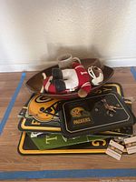 Pile of Packers and Steelers memorabilia including metal signs, a clock, football bowl, plush bear, and mug on a wooden floor near a wall.