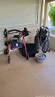 Photo showing red Drive brand walker with front wheels and brakes next to folding wheelchair and small power lifting seat. Grabber stick and blood pressure cuff visible on walker seat.