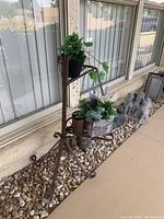 Metal plant stand with multiple potted plants against house exterior wall on stone bed