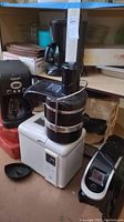 Photo showing multiple kitchen appliances including black Cuisinart coffee maker, black Jack LaLanne juicer, white Toastmaster bread maker and others on shelving.