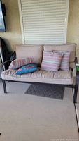 Front view of the metal frame loveseat with beige cushions and multiple pillows including two striped beige/red and one blue pillow.