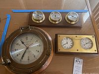 Photo showing 3 brass nautical instruments mounted on wood (two clocks and one barometer) and a large 13-inch Bell Clock Co ship’s clock with porthole casing.