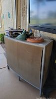 Metal framed outdoor bar on wheels with mesh front panel, green bird yard statue, clay circular object, TV screen in background.