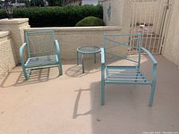 Outdoor patio setting showing two light blue metal chairs and one matching metal round table with glass top arranged on a concrete floor against a beige wall.