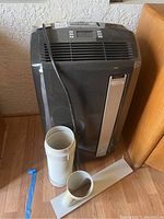 Front view of the DeLonghi portable air conditioner with attached power cord, exhaust hose, and venting panel on the floor next to it.