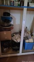 Shelf containing large metal pot, various plastic storage containers with lids, bags possibly with packing materials, and other kitchen items arranged together.