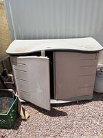 Rubbermaid beige plastic outdoor storage cabinet with left door open showing inside space and some dirt on top surface.