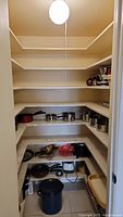 View of shelves with stainless steel pots, red plastic bowls, Crockpot on floor, and other small appliances.