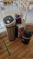 Photo showing three trash bins positioned next to a stair gate inside home, two stainless steel bins one with sensor lid and one with trash bag, plus a smaller black plastic bin with red handle inside