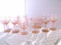 Full view of the set with 4 pink depression glass wine glasses, 2 smaller glasses, pitcher and pedestal dish on white patterned tablecloth