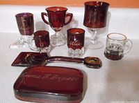 Photo showing assortment of ruby red souvenir glass items including boot, shovel, cup, paperweight, and mugs.