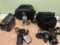 Photo showing all main items including JVC camcorder, Fuji FinePix S camera, two black carrying bags, cables, battery, and memory cards on tile floor with pale green wall background.