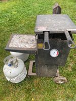 Front and side view of black Cajun deep fryer with side shelf and two fryer baskets, sits on grass next to one white propane tank.