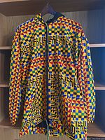 Front view of hooded jacket hanging on a wooden shelf. Vibrant multicolor geometric African print fabric, mostly yellow, red, blue, and green, with full zipper and two front pockets.