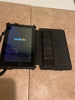 Kindle Fire tablet powered on with screen visible, in black protective folio case, laid open on surface.