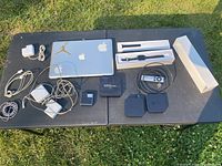 Full lot overview photo showing Apple laptop with Jordan and Apple stickers, two Apple TV units each with visible wear, Apple Watch in box with band, AKASO MXQ PRO 4K media device, iPad mini cover, iPod Nano and multiple power cords and chargers.