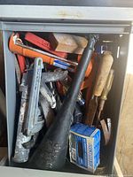 Photo showing large pipe wrench with red handle, metal funnel, multiple chisels with wooden handles, small box of fasteners in metal drawer.