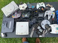 Top-down view of two Sony PlayStation consoles, Sega Dreamcast console, assorted controllers and power/audio cables on a table