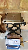 Photo of Cuisinart portable propane BBQ grill with foldable legs and closed lid with logo, placed indoors on cardboard and box.