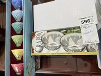 Top view showing boxed set of six clear glass dessert bowls with swirl pattern inside display box and colorful teacups stacked inside box on left side.