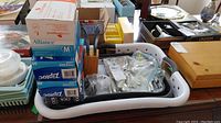 Photo showing a white plastic tray containing assorted cutlery pieces in plastic bags, wooden knife stand with knives, boxes of gloves and Ziploc bags stacked on top, along with a collapsible sink strainer basket beneath