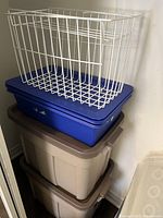 Stack of two large beige storage bins with lids, one blue storage bin with lid, and two white metal wire baskets on top.