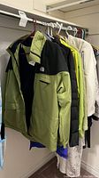 Photo showing nine jackets hanging on a rod including a green and black North Face jacket, neon yellow jacket, black puffer, and white/light-colored jackets.
