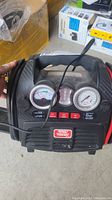 Front view of the jump start and tire inflator charger unit showing pressure and volt gauges, buttons, indicator lights, and battery clamps.