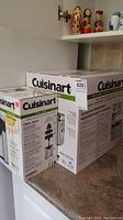 Photo showing boxed Cuisinart bread maker and boxed Cuisinart citrus juicer sitting on kitchen counter next to wall with tiled backsplash and decorative items on shelf.