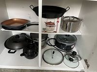 Two shelves filled with assorted pots, pans, and lids including a grill pan, wok, crock pot, and stainless steel pot among others