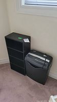 Overall view of the black CD shelf and paper shredder placed side by side against a wall with a window above, positioned on a carpeted floor.