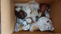 Overhead photo of box filled with various miniature tea set pieces including multiple teapots and cups, some wrapped in bubble wrap and paper.