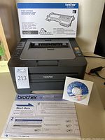Front view of Brother HL-2240 laser printer with extra toner cartridge box, installation CD, and manual papers on table.