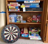Full view of the games and puzzles displayed on a wooden shelf, including board games, puzzles, and a dartboard leaning on the floor