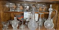 Wide view of all glassware including margarita glasses on upper shelf, cut glass decanters, sugar and creamer, and candy glass decor on lower shelf.