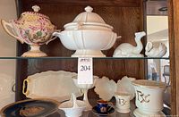 Shelf display showing array of porcelain and ceramic serveware and decorative pieces including urn, tureen, plates, cups, and camel figure