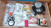 Top view of kitchen drawer displaying small kitchen appliances and utensils including handheld mixer, immersion blender, steamer basket with lid, coffee mug warmer, garlic baker, and gravy separator.