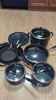 Full set of four Circulon Commercial pots with lids and four matching frying pans, all black with glass lids and metal trim, plus one RevereWare two-handled pot with lid