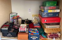 Photo showing a large assortment of games, puzzles, playing cards, and an automatic card shuffler, including a Piatnik giant size playing cards box and several game boxes stacked.