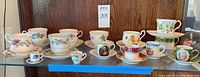 Full display of all 8 teacups and matching saucers arranged on shelf, showing variety of floral and decorative patterns, plus two small souvenir cups and one isolated saucer.