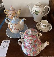 Four decorative teapots with their matching cups displayed side by side on a wooden surface, showing varied floral and bird designs in different colors and styles.
