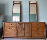 Front view of vintage Thomasville wooden dresser with six drawers on each side and a central cabinet with two doors. Two tall rectangular mirrors with inset tile accents at top of frames are placed on top of dresser.