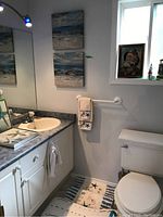 Bathroom scene showing towels with starfish embroidery on towel rod, nautical framed art above toilet, lighthouse figurines on windowsill, mirror reflection of room and sink.
